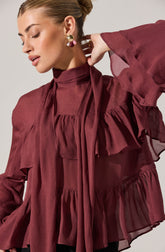 A woman is wearing a dark red, semi-sheer blouse with a high neckline. The blouse features ruffled details around the shoulders and chest, with ruffles extending down the front and along the sleeves. The fabric appears lightweight and flowy, creating a soft and feminine look. She is accessorizing with large, ornate earrings with gold, pearl, and gemstone elements.